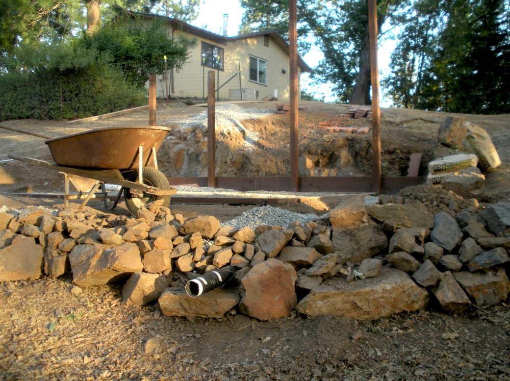 A close up shot of a poured greenhouse foundation on a stone base with drainage and wall posts up and a wheel barrow to the left.