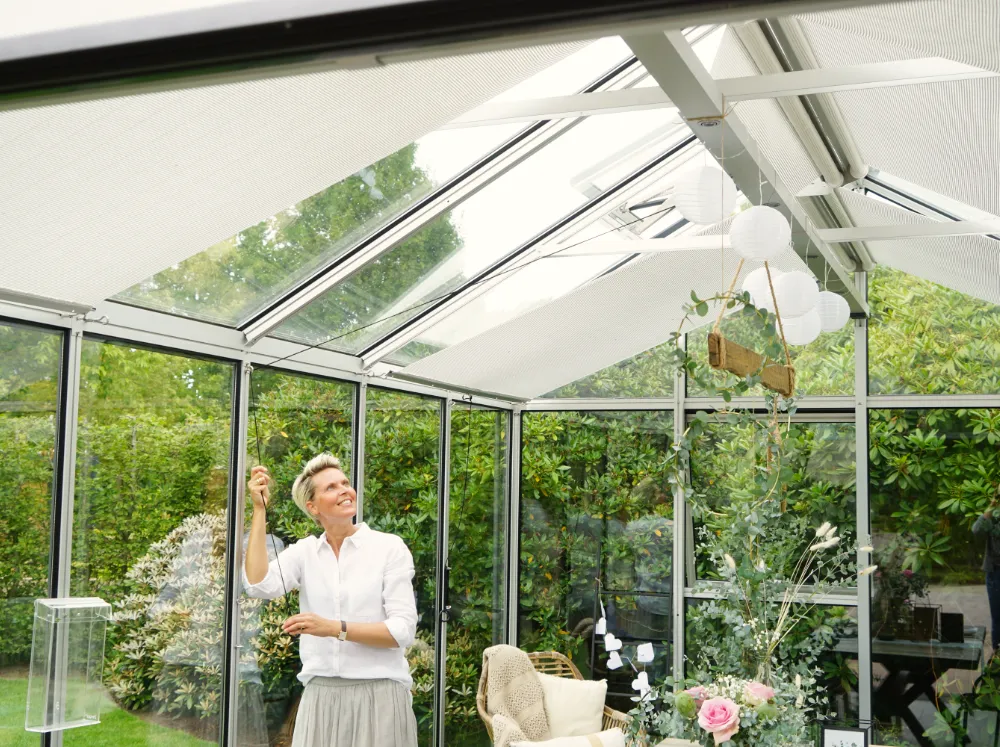 Retractable Roll-up Shade Curtains installed in a glass greenhouse with a woman adjusting them
